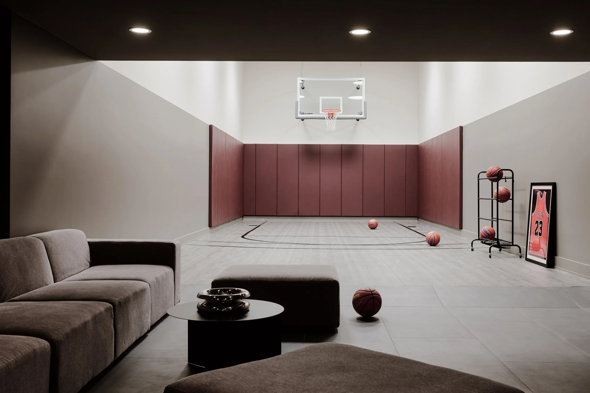 A home basketball court with a hoop, basketballs on the floor and shelves, and framed sports memorabilia, with seating area in foreground.