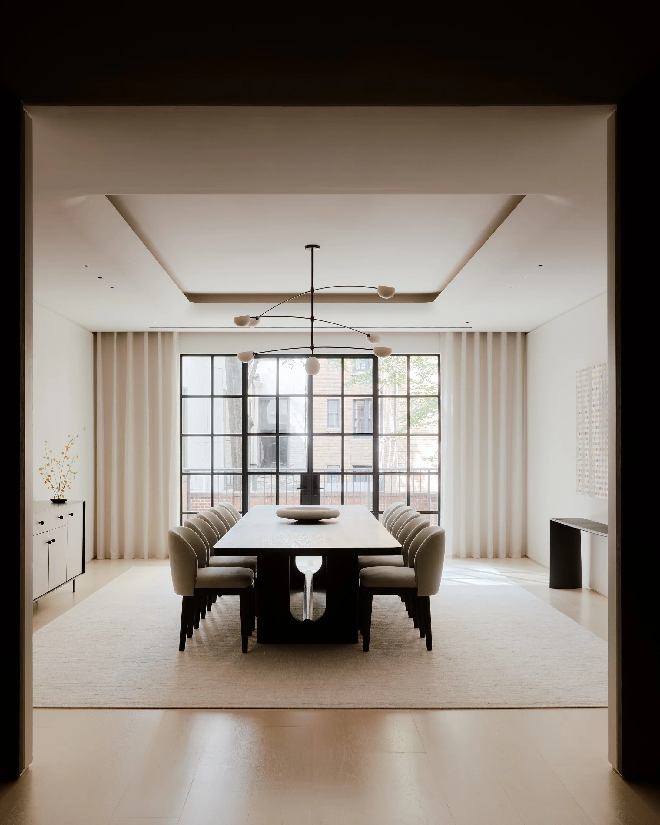 A modern dining room with a large black rectangular table surrounded by eight beige upholstered chairs. The room features a large window with grid panes, white curtains, and a built-in cabinet with a potted plant. A contemporary chandelier hangs above the table, and a neutral-toned rug covers the floor.