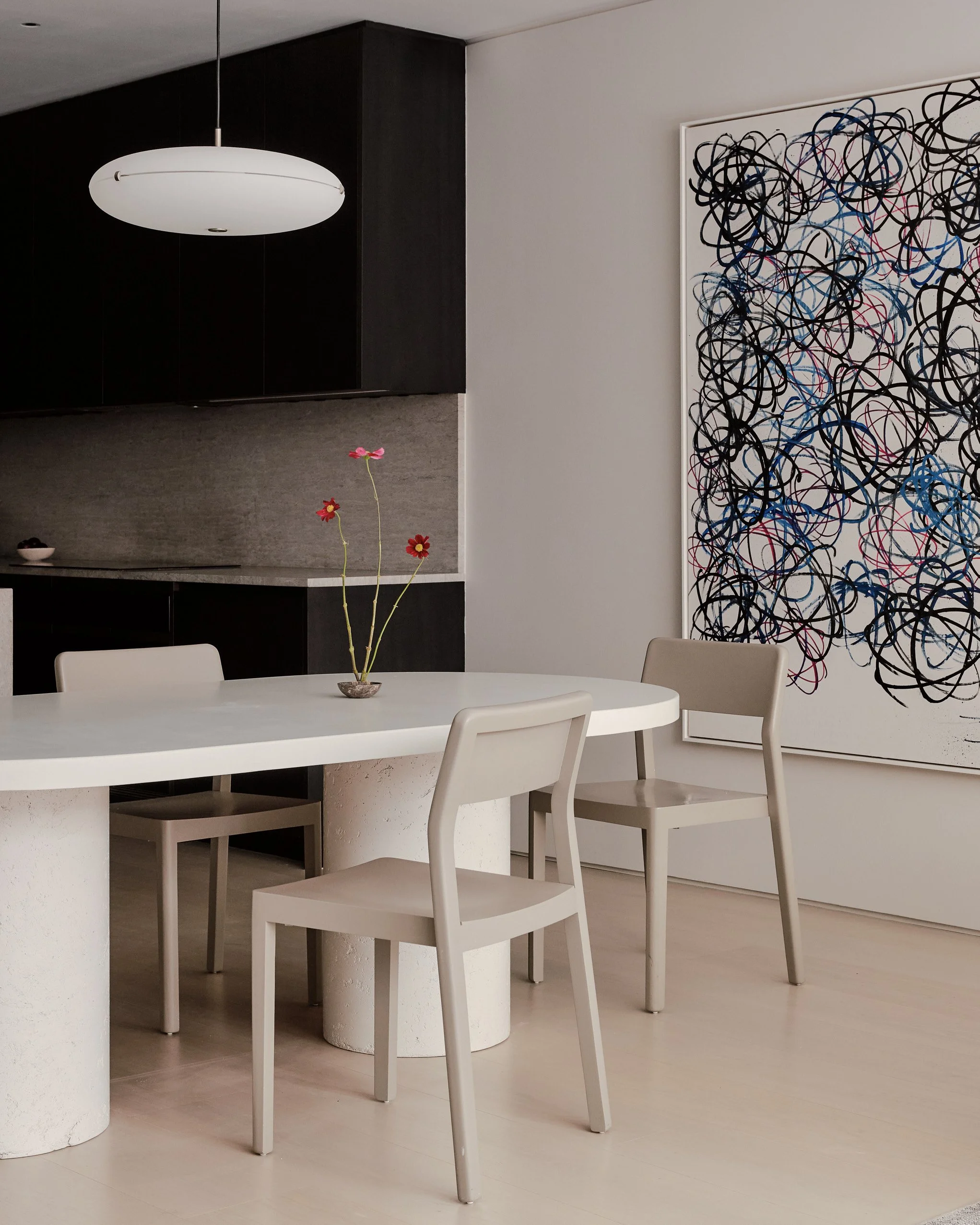 A white dining table with four matching chairs, a small vase with pink and red flowers on the table, a large black-and-white abstract painting on the wall, and a ceiling light fixture in a minimalist modern interior.