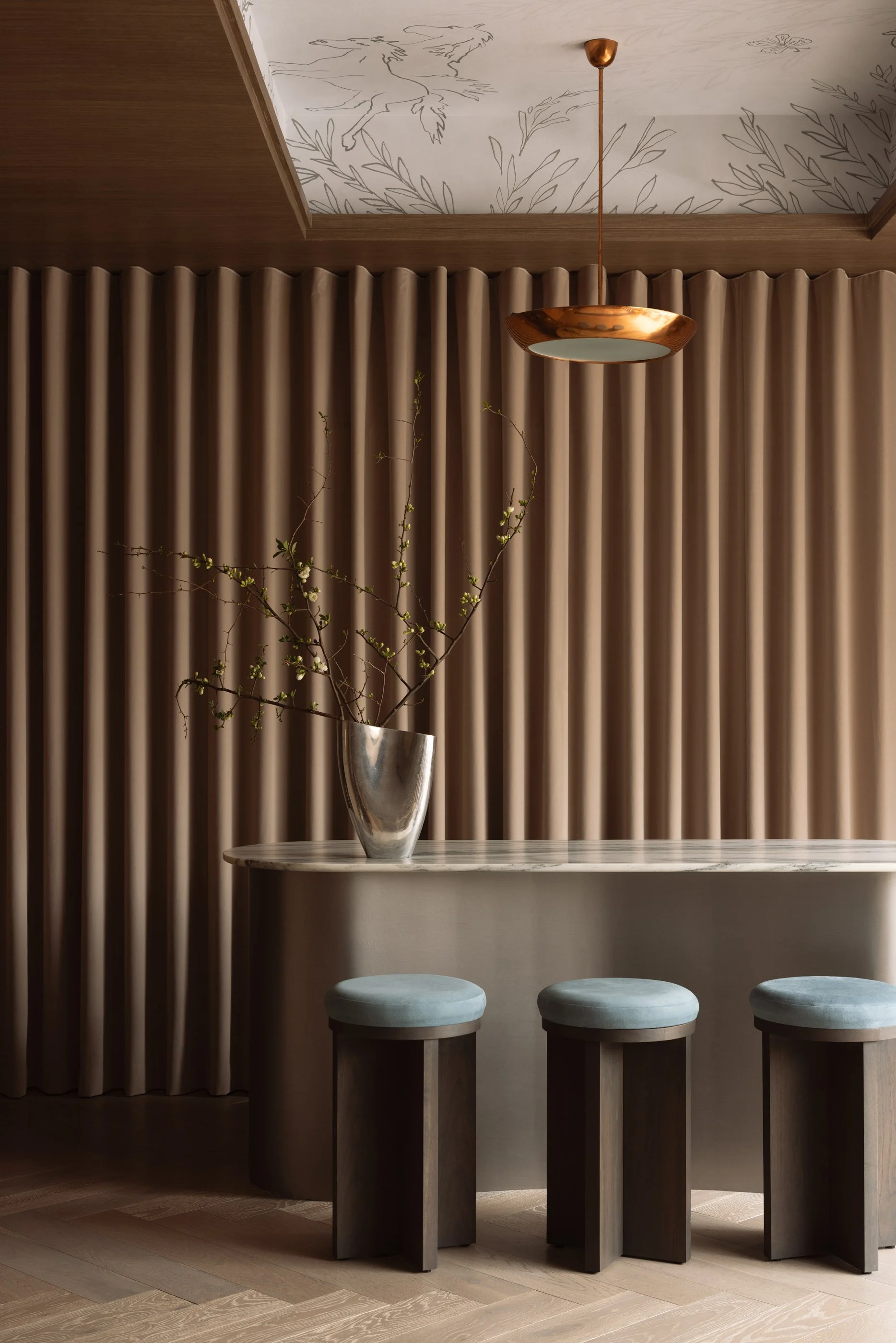 Interior design with beige barstools lined up at a marble-topped bar, a small vase with branch and leaves, beige drapes, and a ceiling with artistic line drawings of animals and plants.