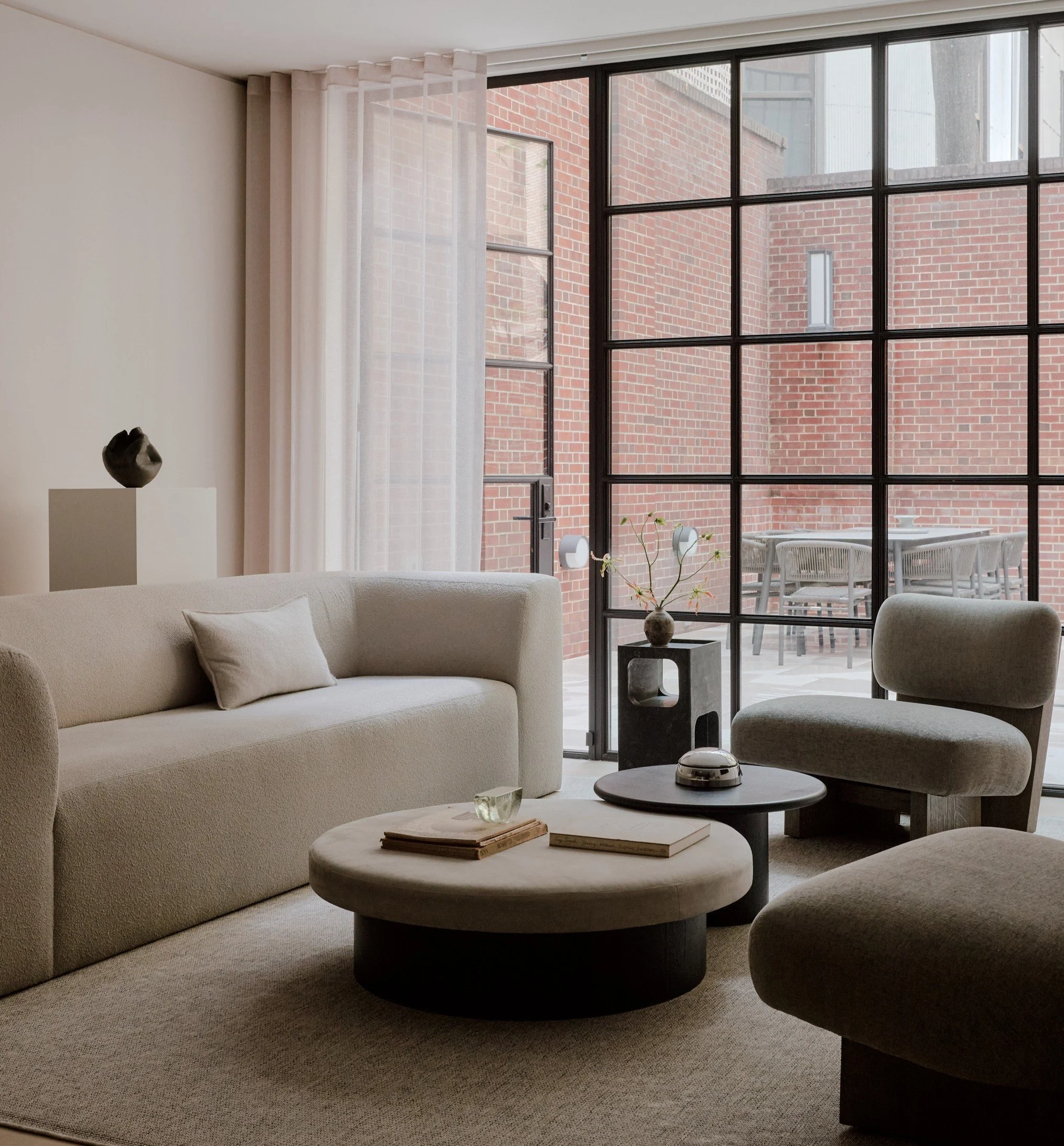 Living room with beige sofa, matching armchair, round coffee table with books and a decorative object, large floor-to-ceiling window with a balcony and brick building outside, potted plant on the table.