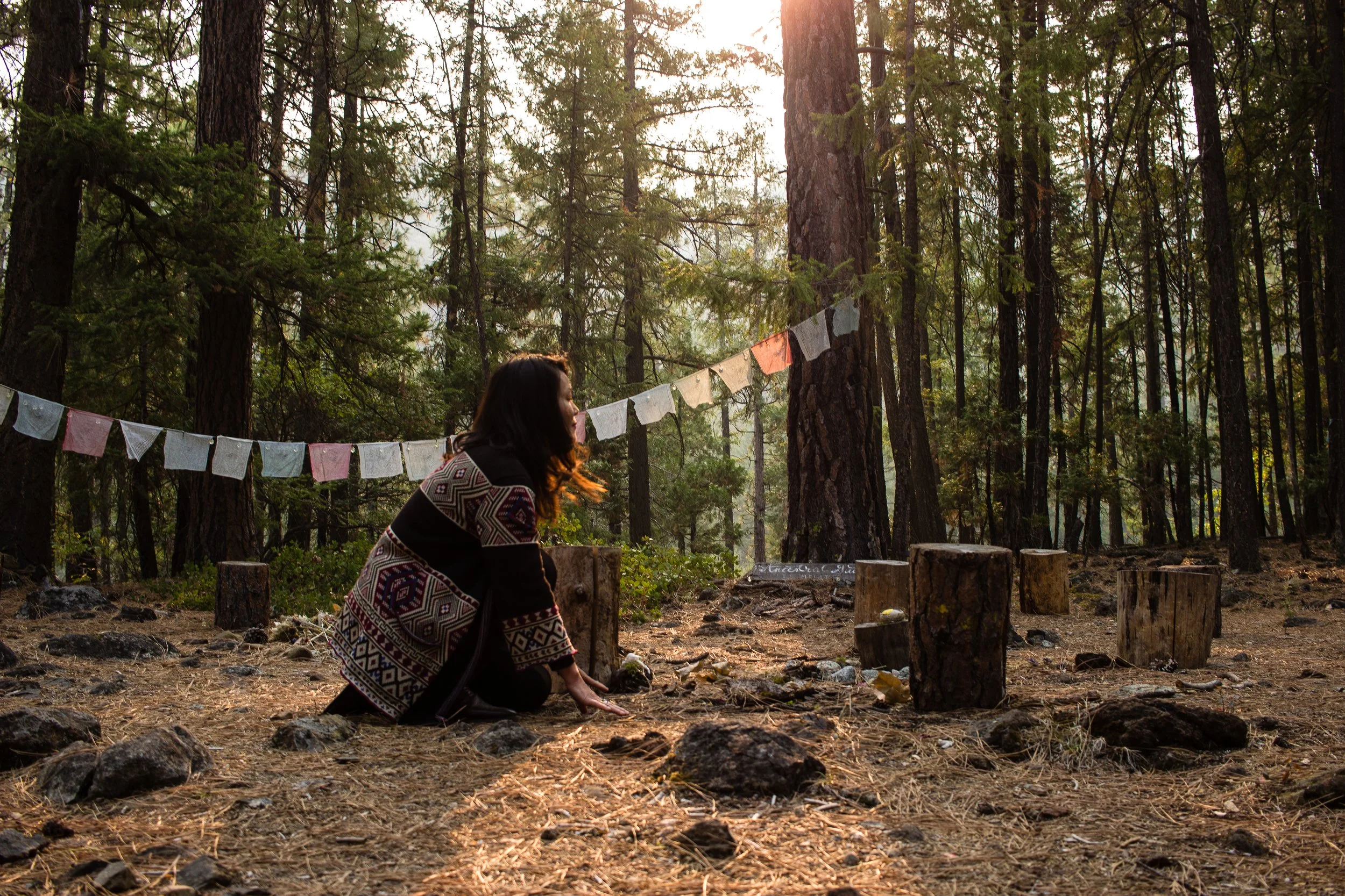 Woman kneeling in a ceremony circle strung with prayer flags at Hestia's Mount Shasta forest sanctuary, California