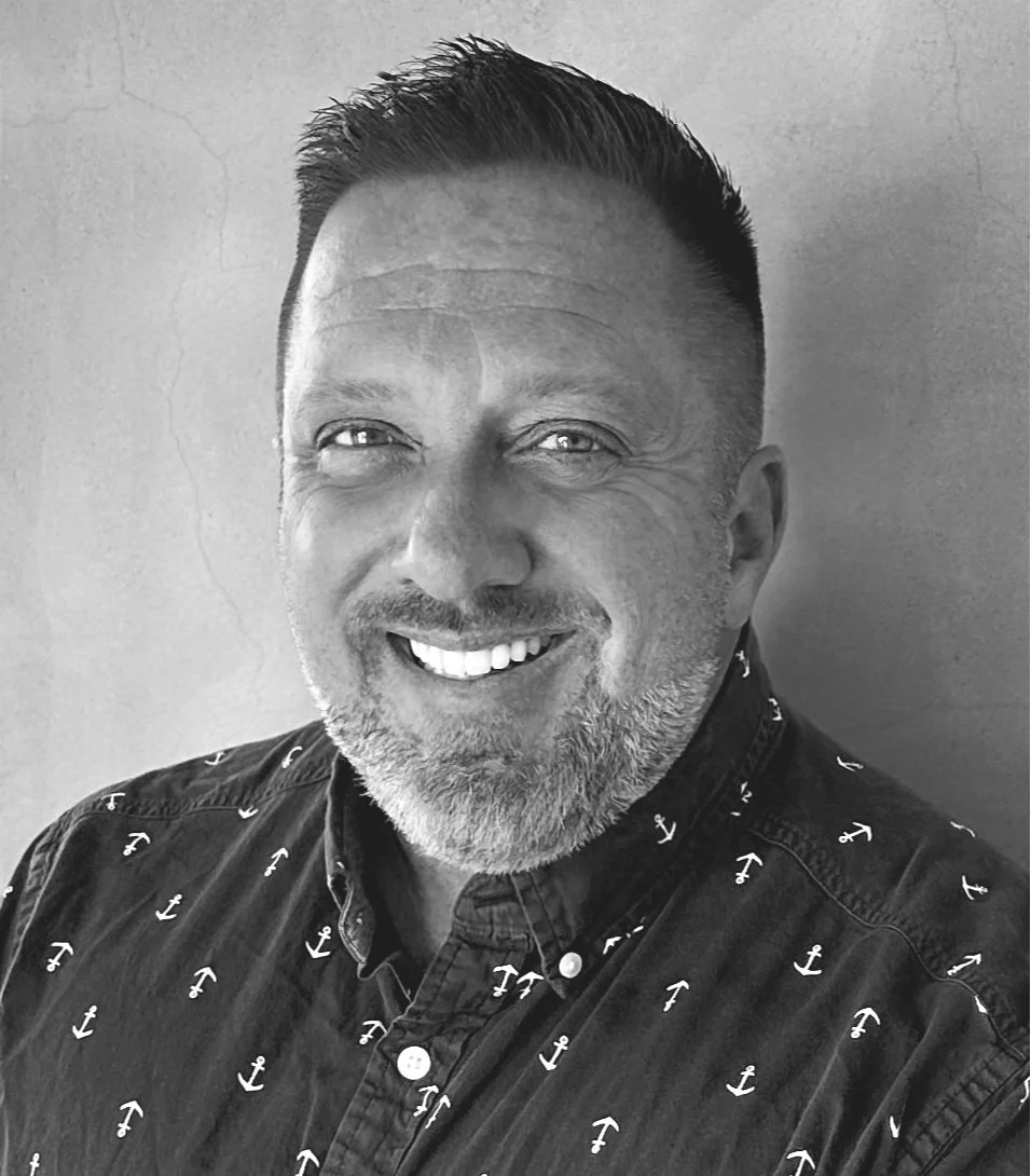 Black and white photo of smiling man with short hair, facial hair, wearing a button-up shirt with small anchor pattern, standing against a plain wall.