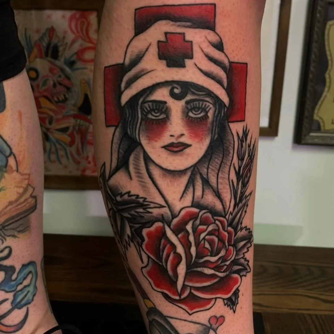 Take a peek at this traditional style nurse by Zack! Tattooed in soft, muted tones paired with strong red eye makeup and bold blackwork. She is framed by wheat stalks and other vegetation, with a central red rose resting beneath her bust. A large red