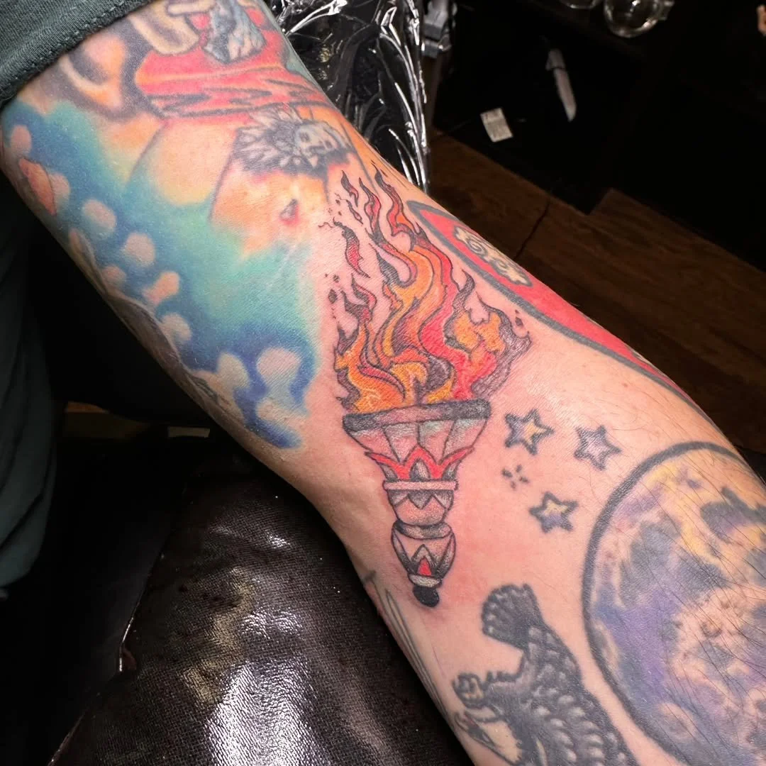 Check out this blazing ceremonial torch tattoo for Chris! Featuring fiery red, orange, and yellow flames rising from a detailed base, this vibrant filler fits right in alongside the surrounding stars and colorful sleeve work. 🌟🔮🖤

Whether you need