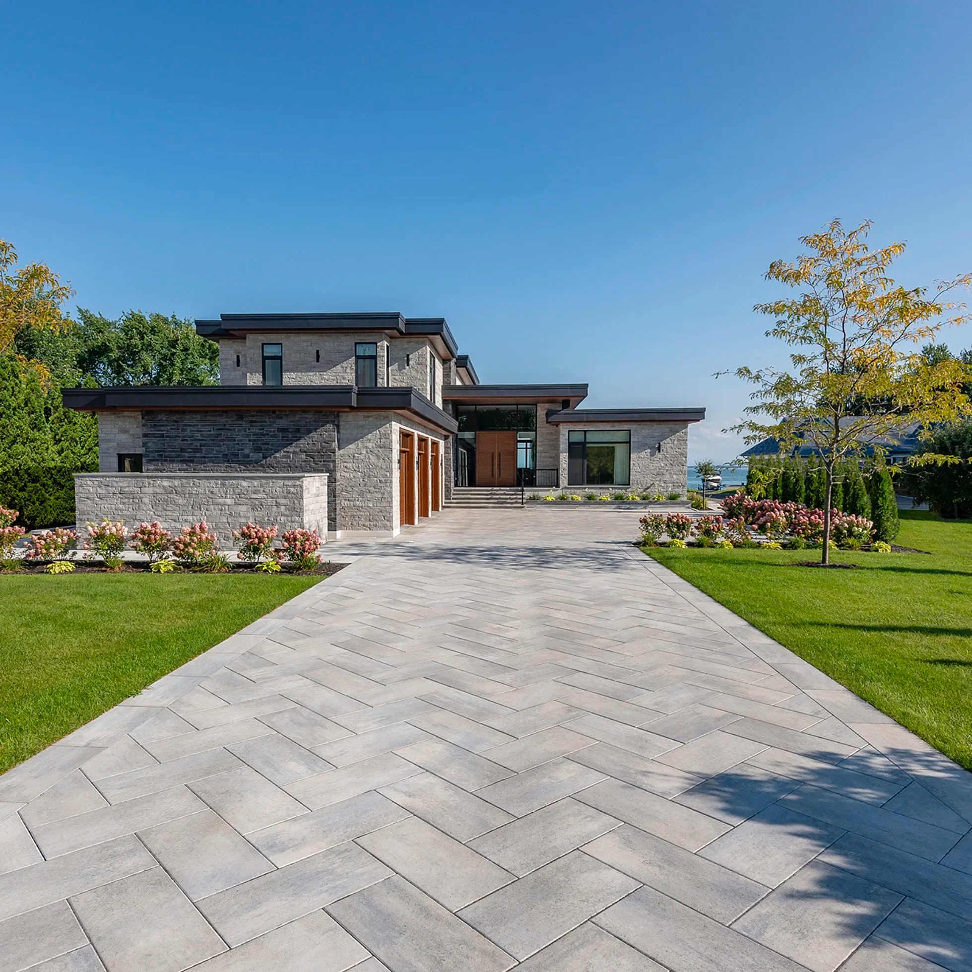 Modern house with a stone and wood exterior, surrounded by a well-manicured lawn, colorful flowers, and trees, with a paved pathway leading to the entrance under a clear blue sky.