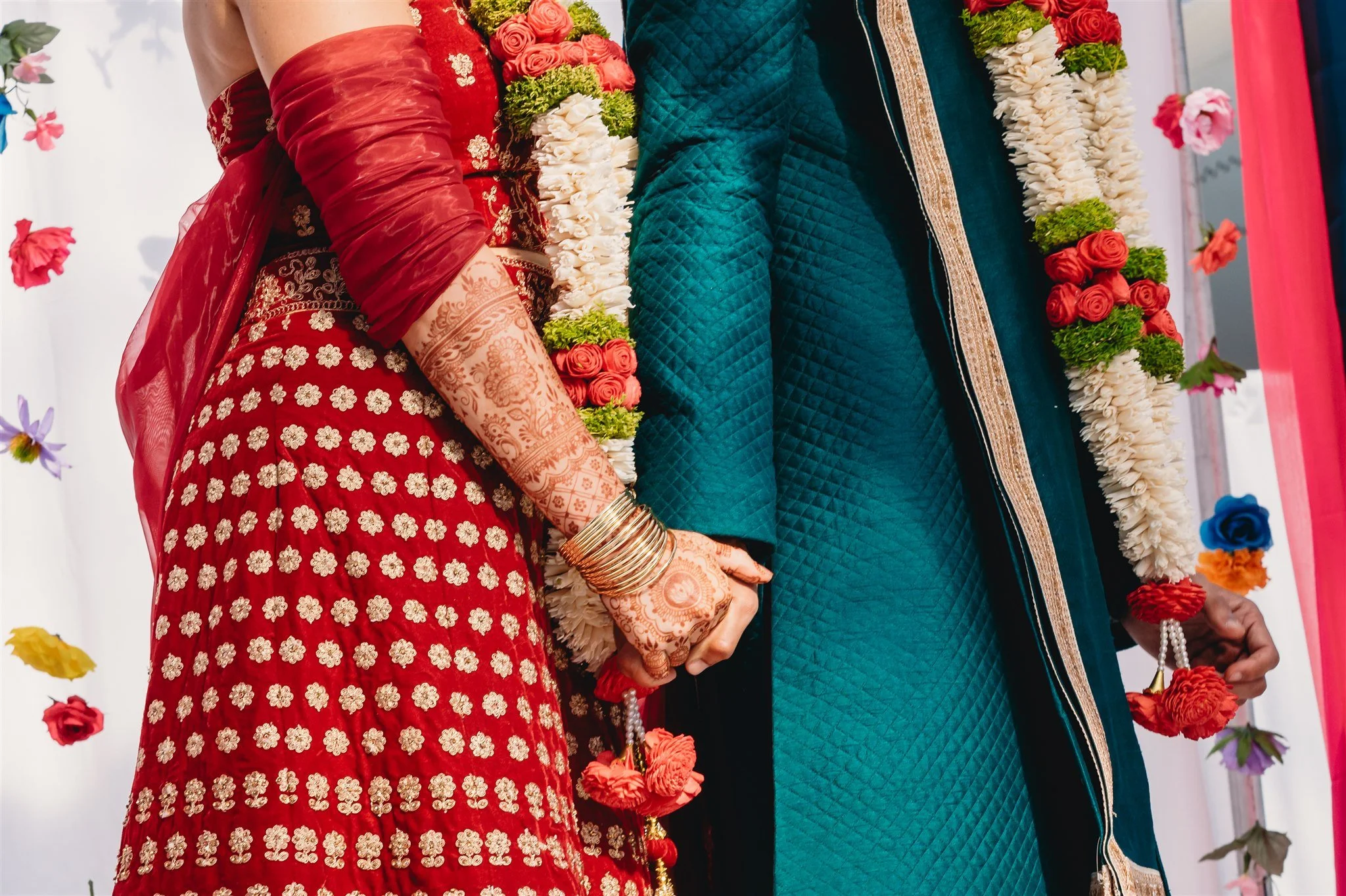 A bride and groom holding hands at an Indian wedding ceremony. The bride is dressed in a red saree with floral embroidery and traditional jewelry. The groom is wearing a teal kurta with a matching dupatta. Both are wearing flower garlands around thei
