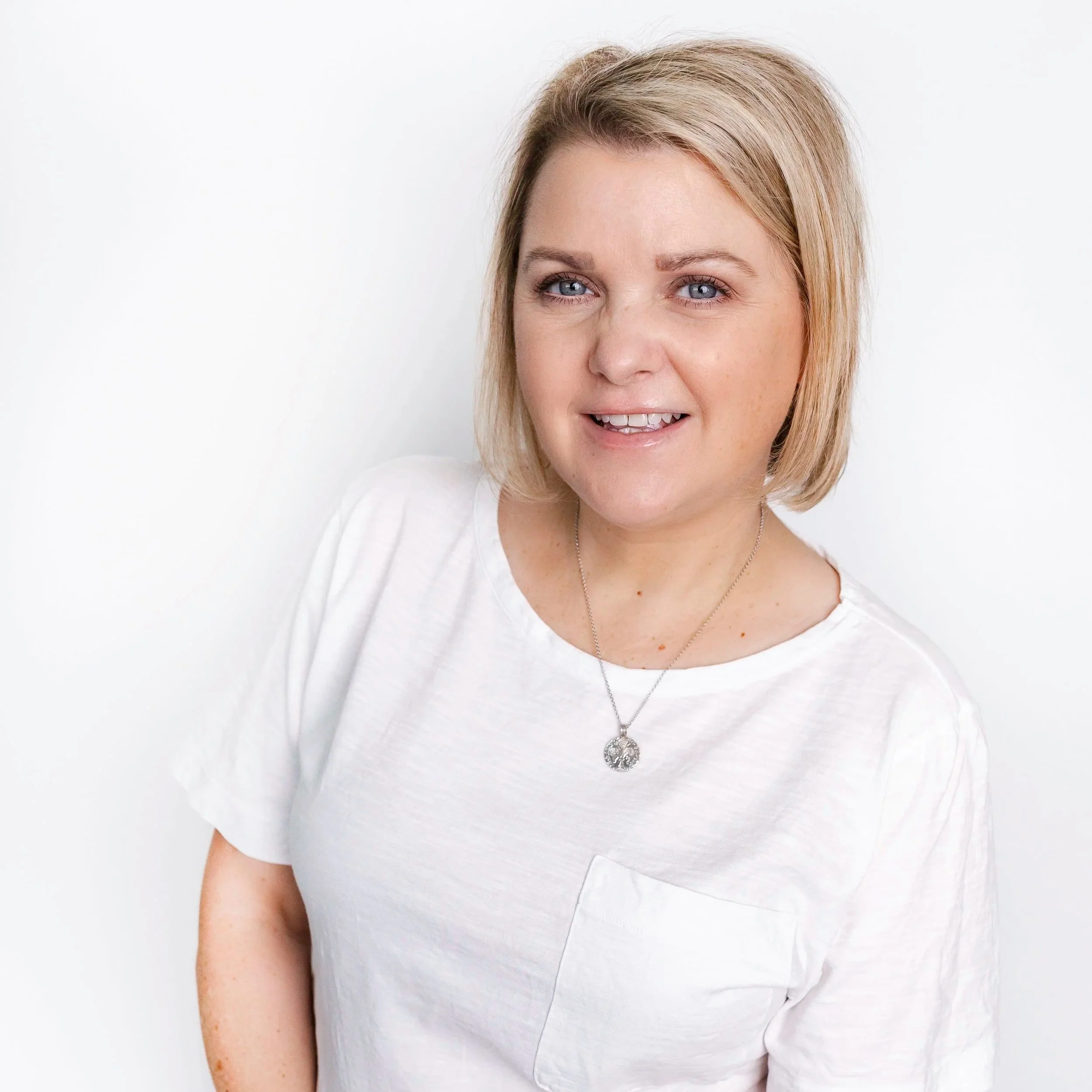 Headshot of a woman with blonde hair, blue eyes, wearing a white shirt and a silver necklace with a pendant, smiling slightly against a plain white background.