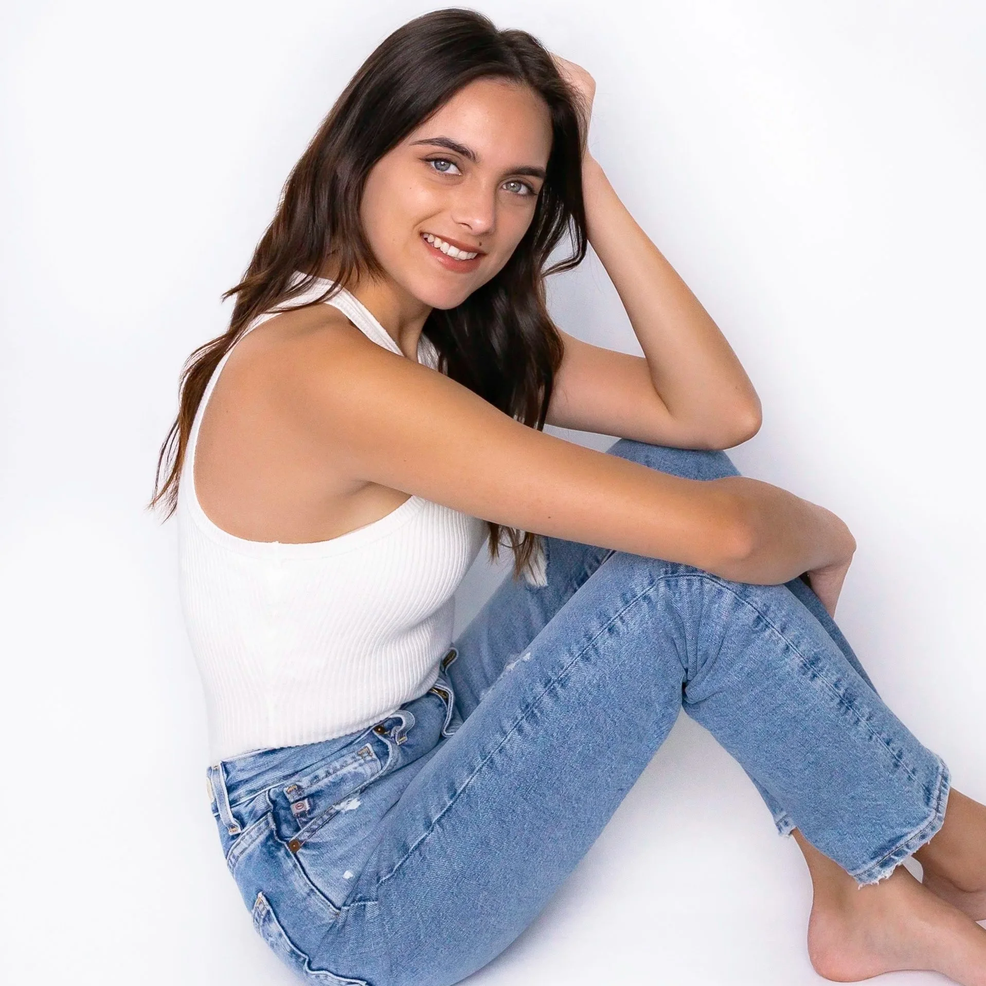 A young woman with long brown hair, wearing a white tank top and blue jeans, sitting barefoot against a white background, smiling at the camera.
