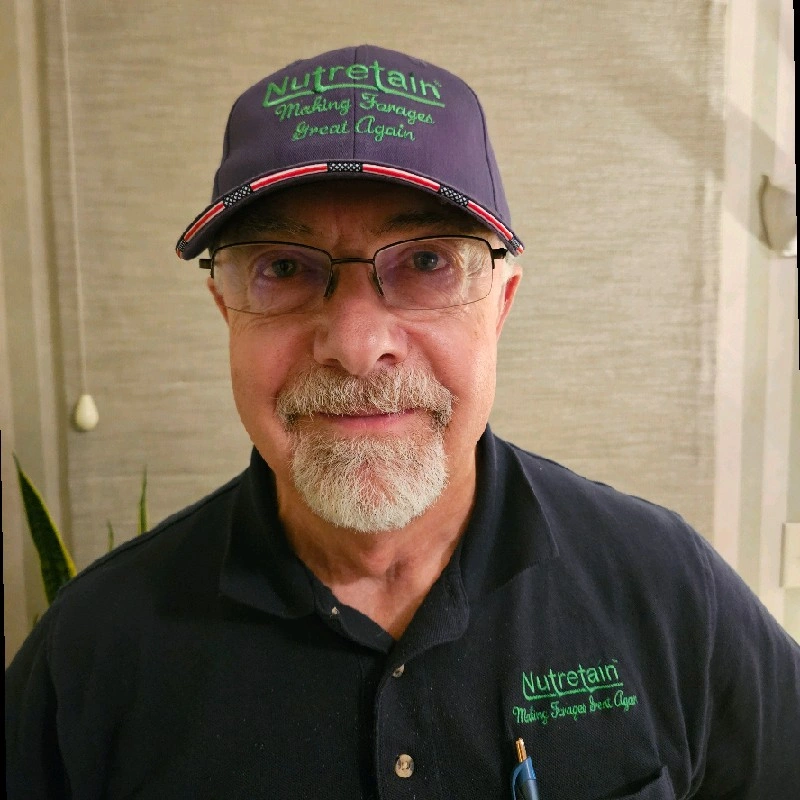 A smiling older man with glasses, a white beard, and a mustache wearing a navy blue polo shirt and a navy cap with green and purple embroidery. The cap and shirt both display the logo and slogan of Nutretain, 'Making Strange Beings Great Again.' he is indoors with a textured beige wall and a potted plant behind him.