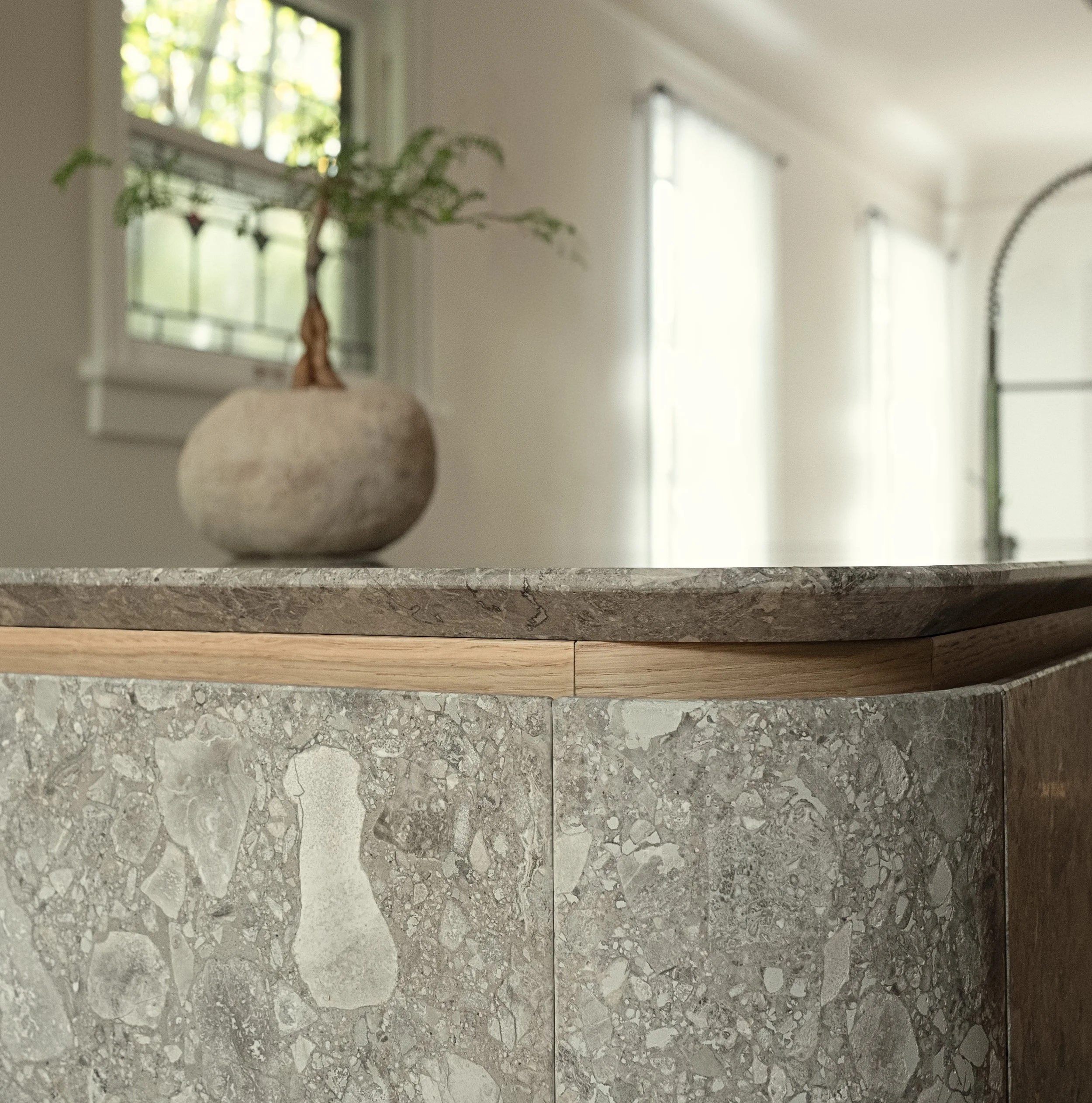 Dettaglio di un bancone in pietra con dettaglio in legno e una pianta in un vaso di pietra, con ambientazione luminosa e finestra con tende in una stanza moderna.