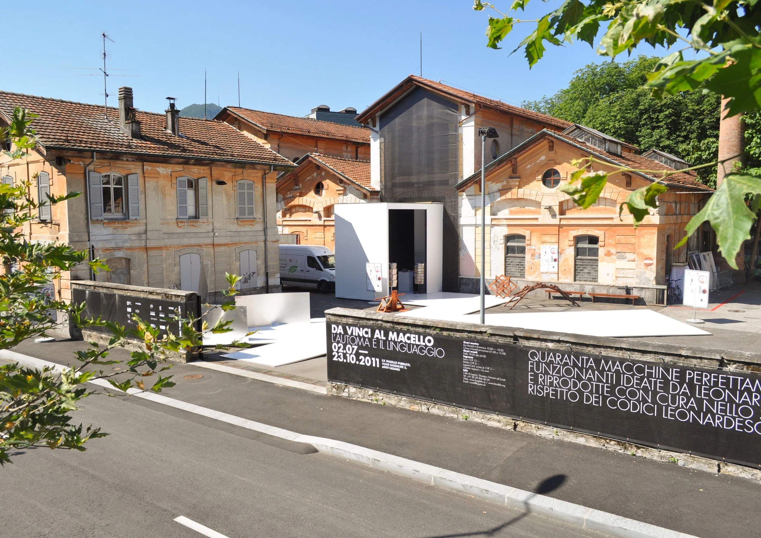 Mostra artistica all'aperto con una piattaforma bianca e un edificio storico alle spalle, con cartelloni informativi e strutture temporanee, in un'area urbana con edifici in stile classico e alcune piante.