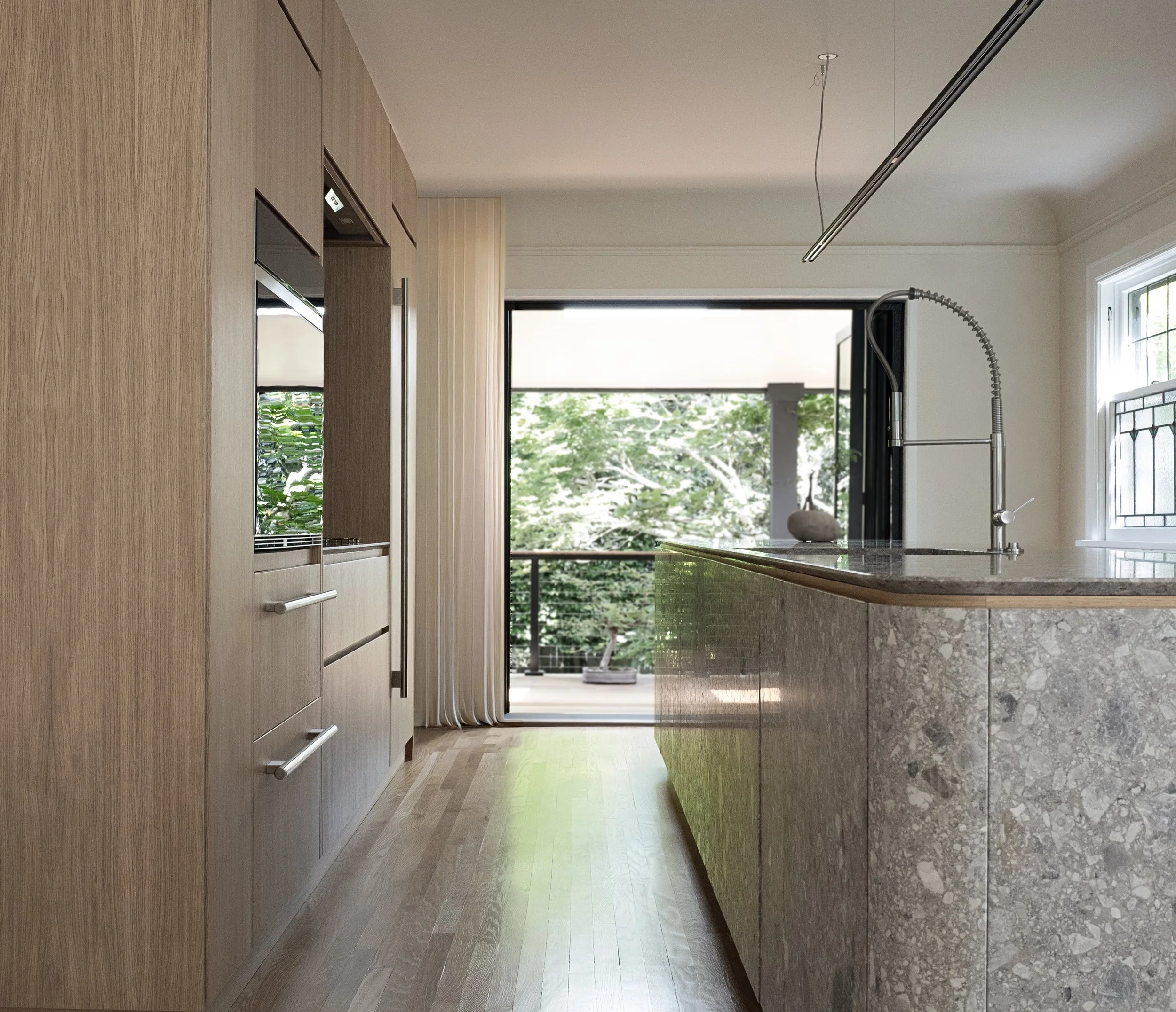 Interno di una cucina moderna con parete in legno chiaro, piano di lavoro in pietra e grande finestra che si apre su un balcone con vista su alberi verdi.