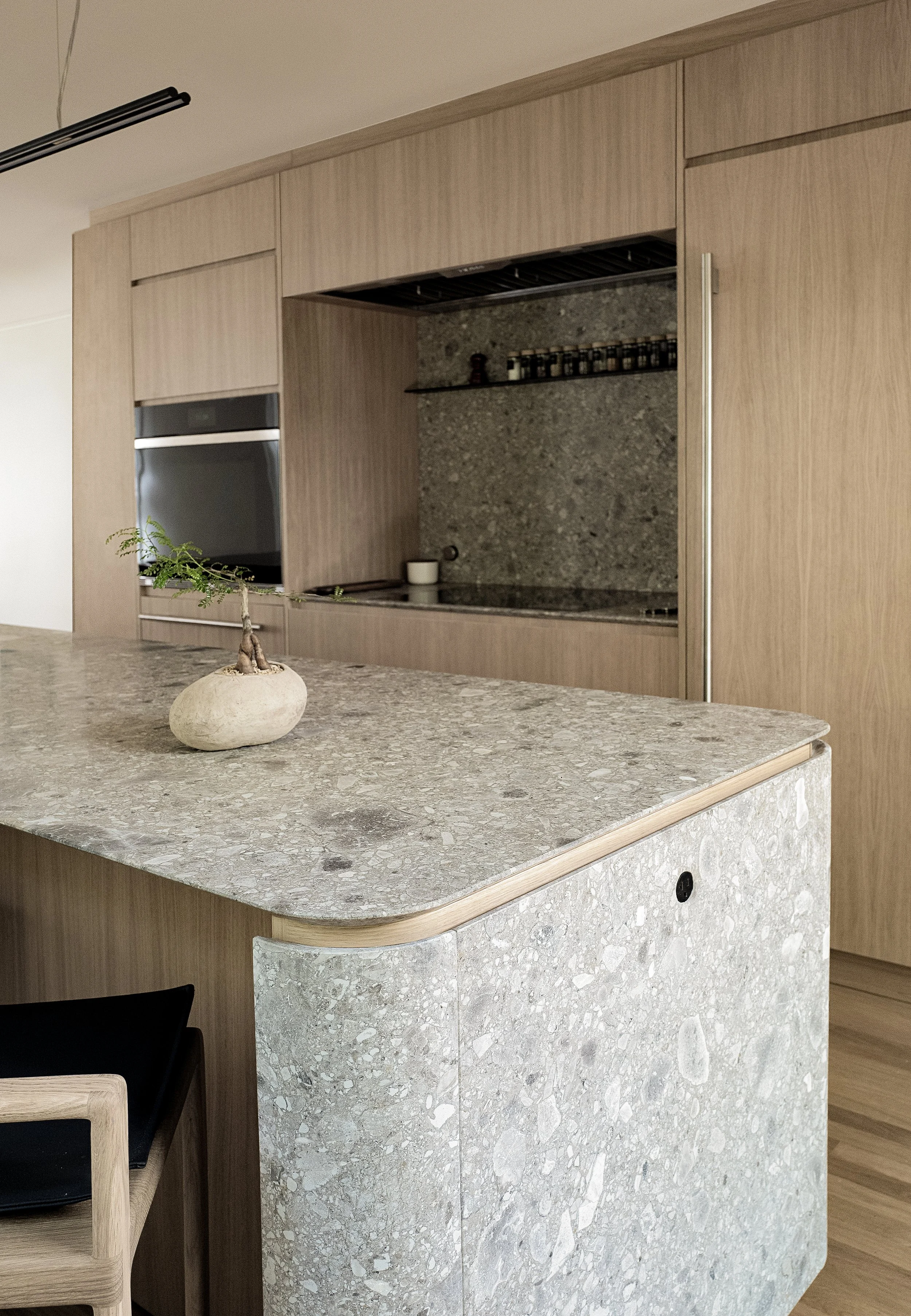 Cucina moderna con piano in pietra, mensola con spezie, ante in legno chiaro, e pianta decorativa su isola in pietra.