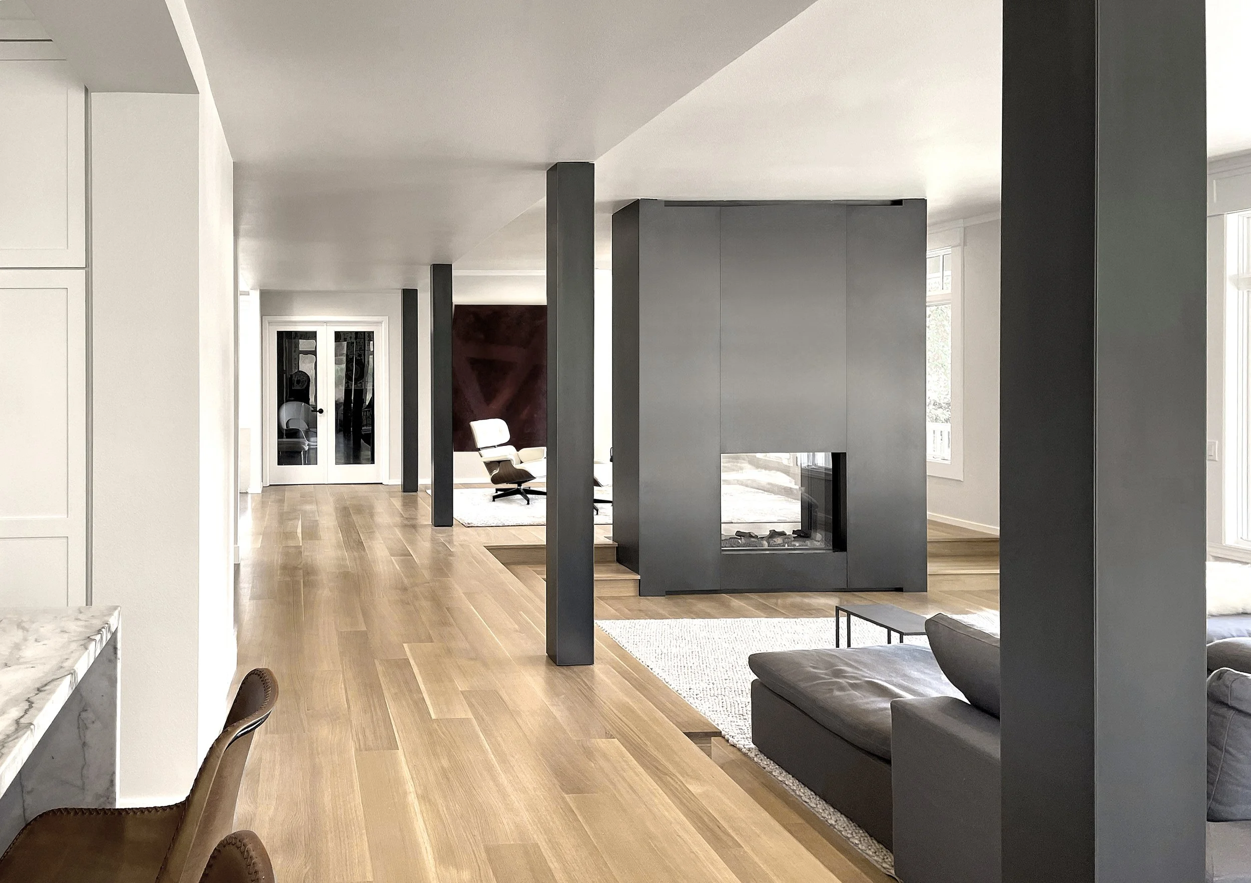 Interno di un appartamento moderno con pavimento in legno chiaro, pareti bianche e una parete divisoria nera con camino. Ci sono divani e una sedia in stile vintage e grandi finestre che lasciano entrare molta luce naturale.