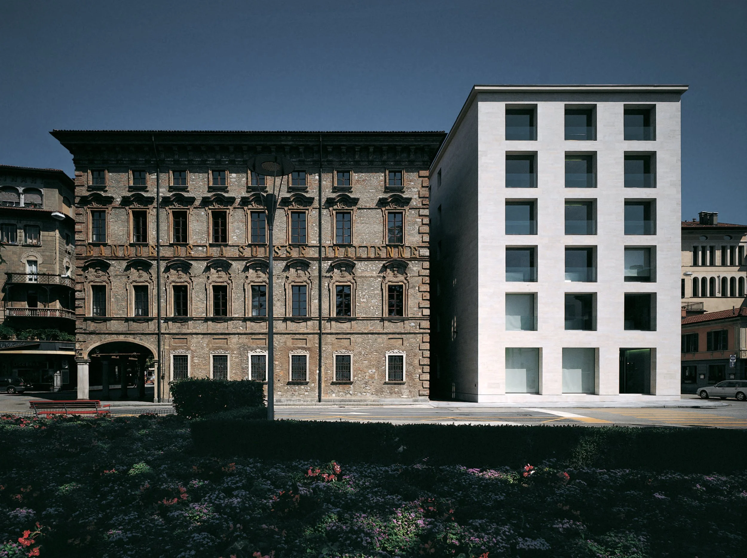 Confronto tra un edificio storico in pietra e un edificio moderno con finestra a nastro. La scena è illuminata dal sole e ci sono piante e fiori sul primo piano.