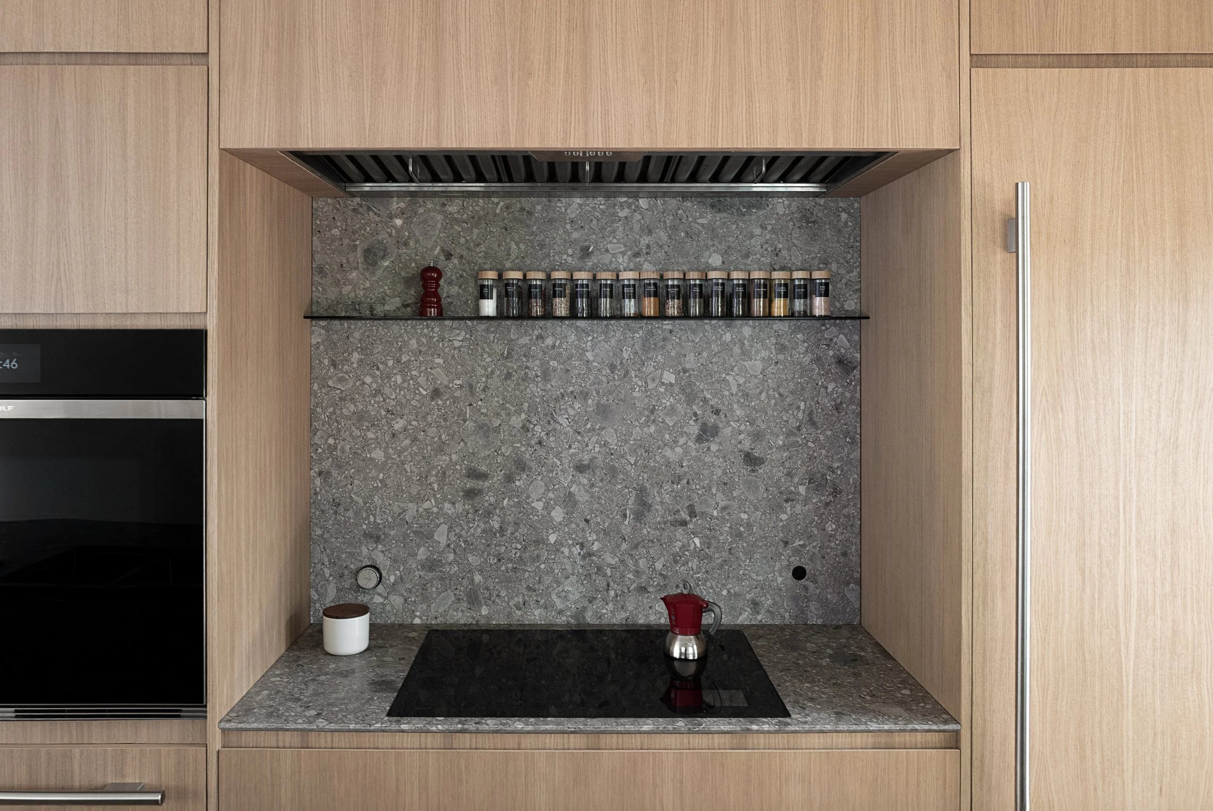 Cucina moderna con ante in legno, piano di lavoro in granito, piani di cottura a induzione, piccolo elettrodomestico rosso, spezie in contenitori trasparenti con coperchio di legno, vicino a parete di marmo grigia.