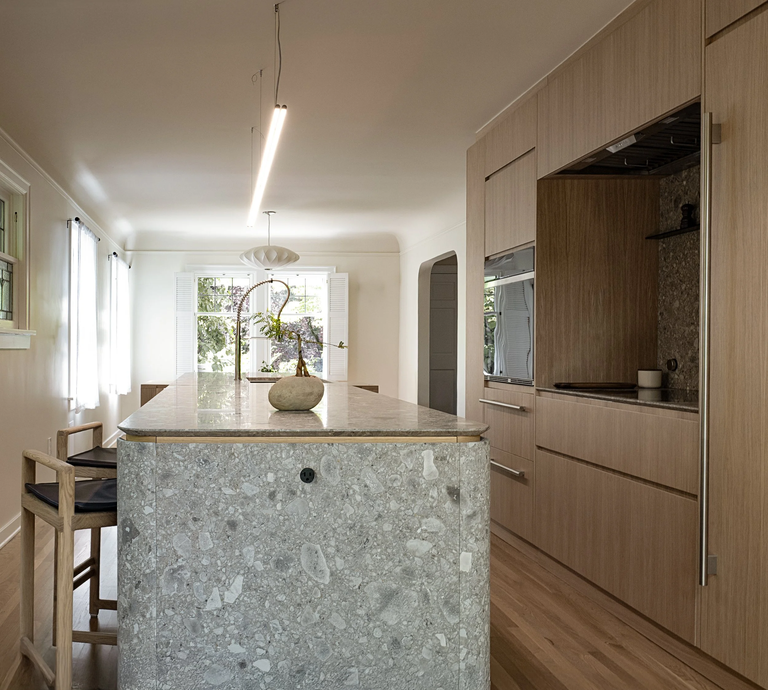 Cucina moderna con isola in pietra, armadi in legno chiaro, grandi finestre con tende bianche, al centro un lavandino con un vaso decorativo.