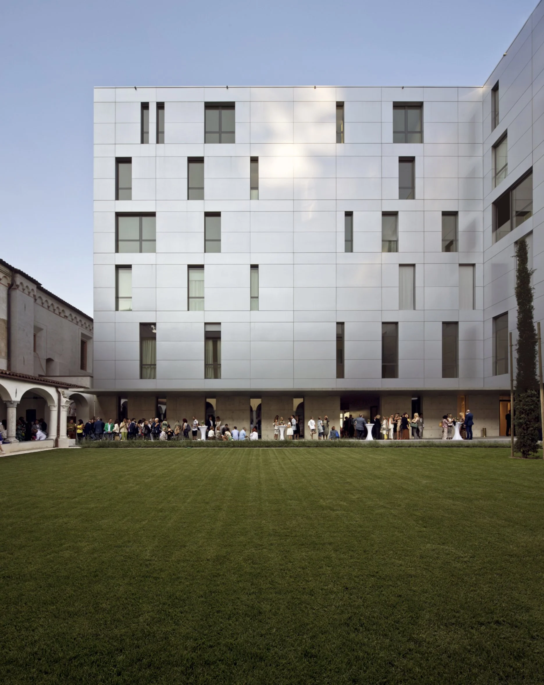 Edificio moderno bianco con molte finestre, persone che partecipano a un evento all'aperto sulla loggia al piano terra, con prato verde in primo piano.