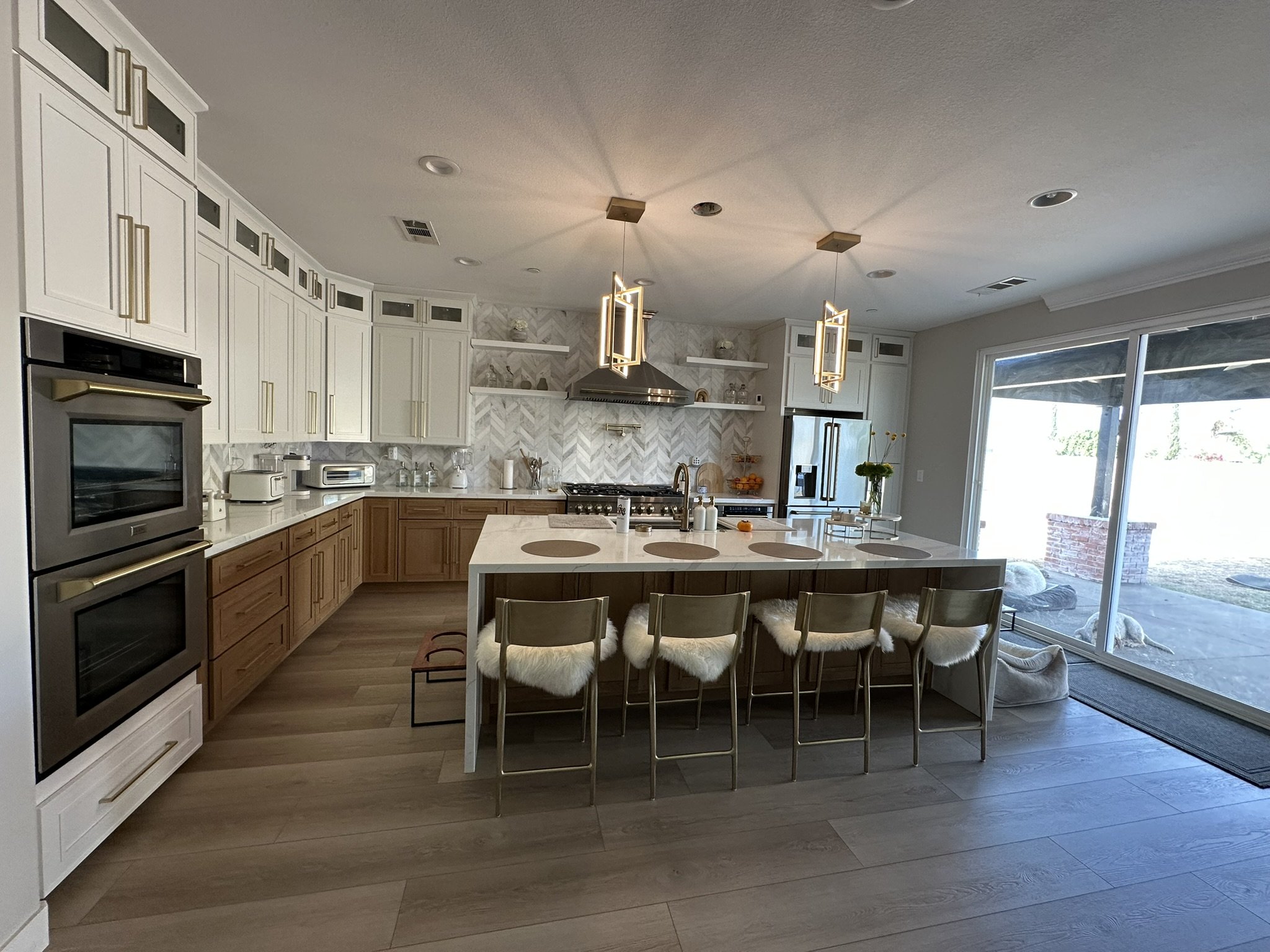 Modern kitchen interior with white cabinets, stainless steel double ovens, island with bar stools, pendant lighting, and large windows opening to a patio.