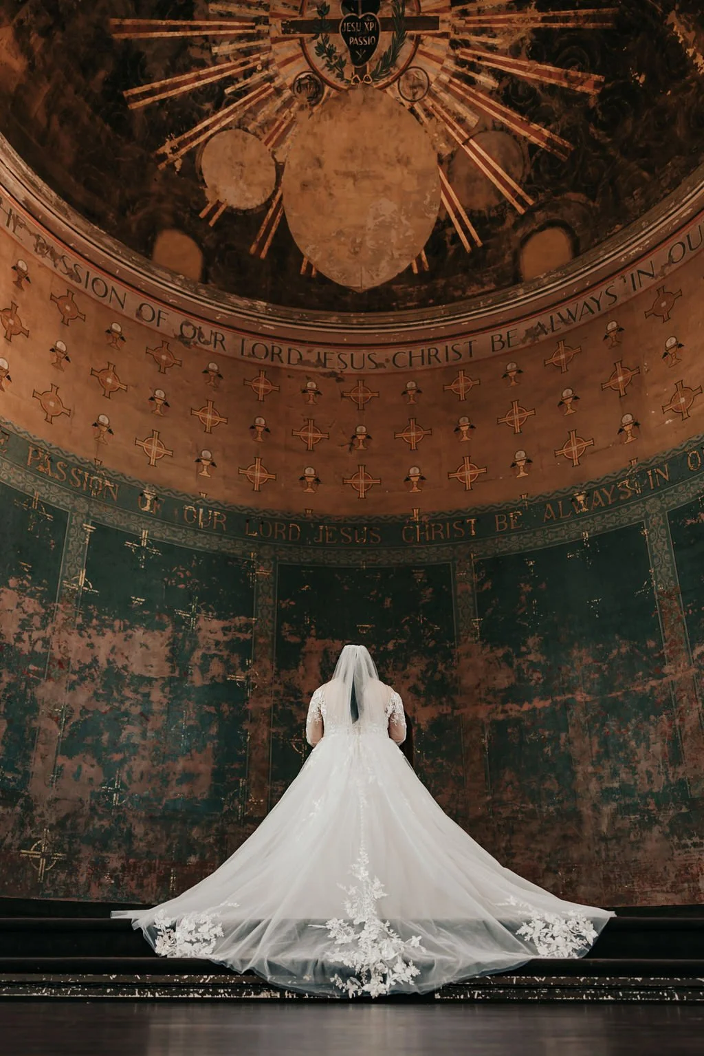 A bride in a white wedding dress with a long train stands in a church, facing a decorative wall with religious text and symbols.