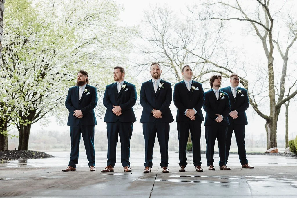 Groom and groomsmen in suits standing outdoors on a cloudy day with trees in the background.