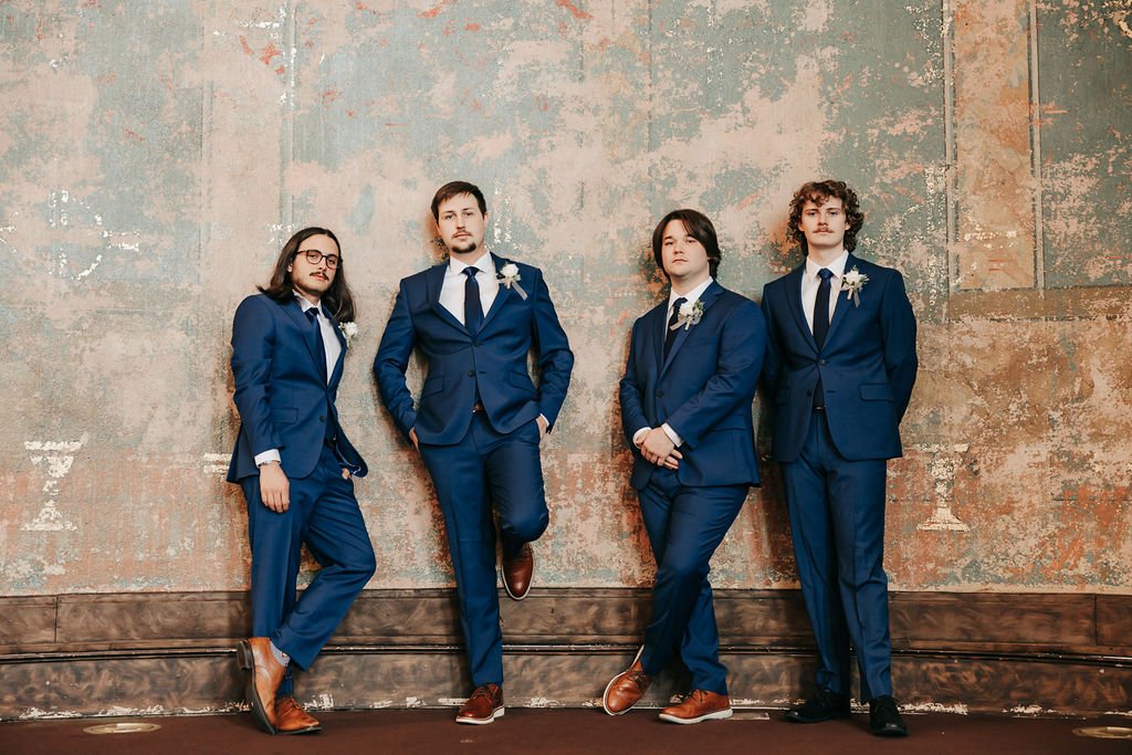Four men in matching blue suits with white shirts, dark ties, and boutonnières, standing against a textured wall, posing confidently.