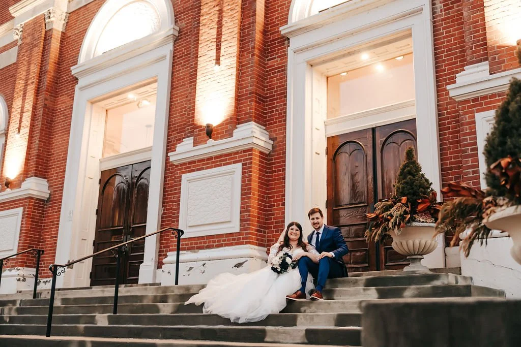 A newlywed couple sitting on the steps of a brick building, posing for a wedding photo. The bride is wearing a white wedding dress and holding a bouquet, and the groom is wearing a blue suit.