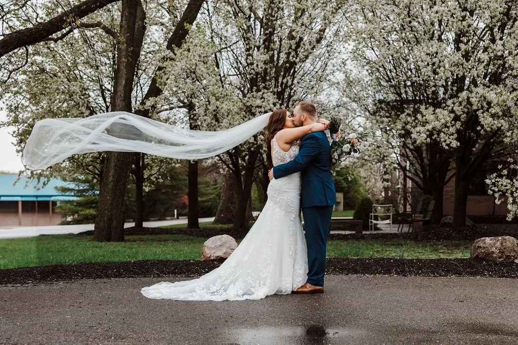 Bride and groom sharing a kiss outdoors, with blossoming trees in the background, the bride wearing a white wedding dress with a veil flowing in the air, and the groom in a blue suit holding a bouquet.