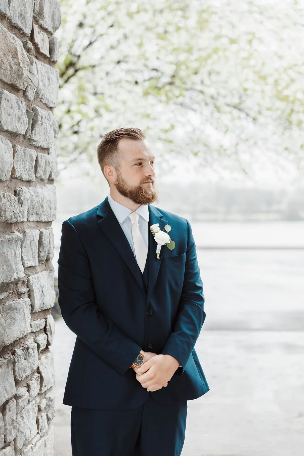 A man in a dark suit with a white boutonniere, standing outdoors near a stone wall with a river and trees in the background.
