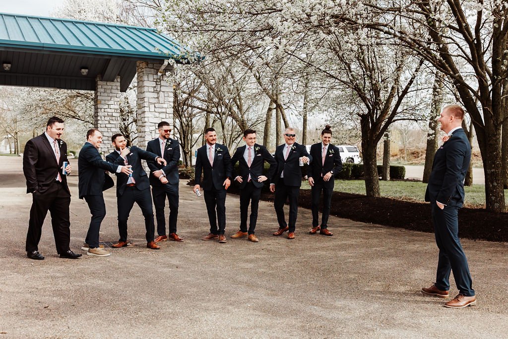 A groom facing his groomsmen, who are dressed in suits, outdoors on a cloudy day near trees and a stone structure with a blue roof.