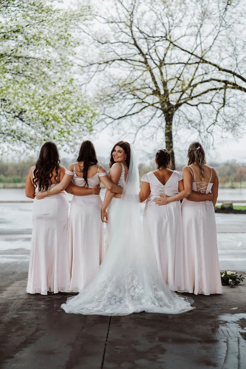 Bride and four bridesmaids in white dresses standing outdoors near a lake, with trees in the background. The bride is smiling and looking over her shoulder.