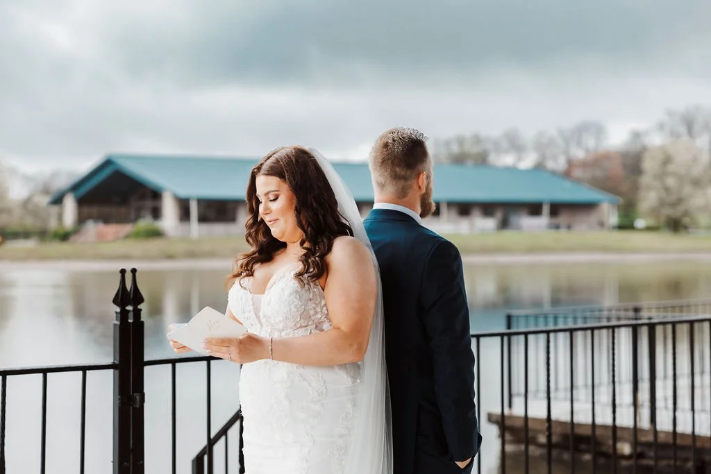 Bride and groom standing back to back near a lake, with the bride reading from a piece of paper, on a cloudy day.