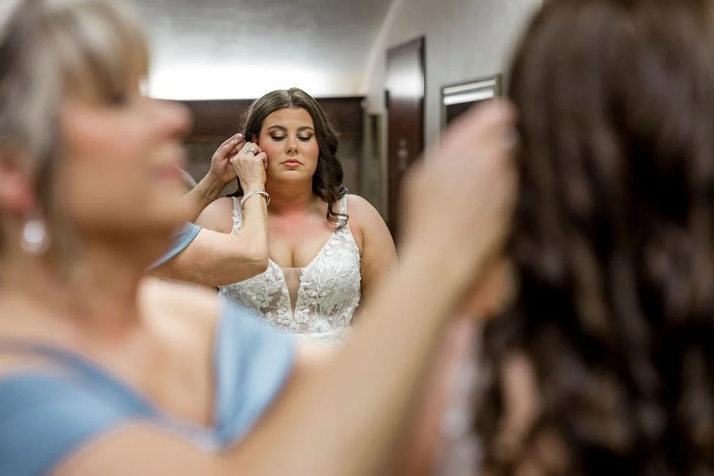 A woman in a white lace dress is being assisted with earrings, with a mirror reflecting her face and makeup.
