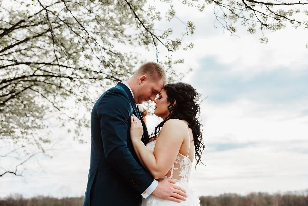 A couple dressed in wedding attire standing close together outdoors with trees and cloudy sky in the background.