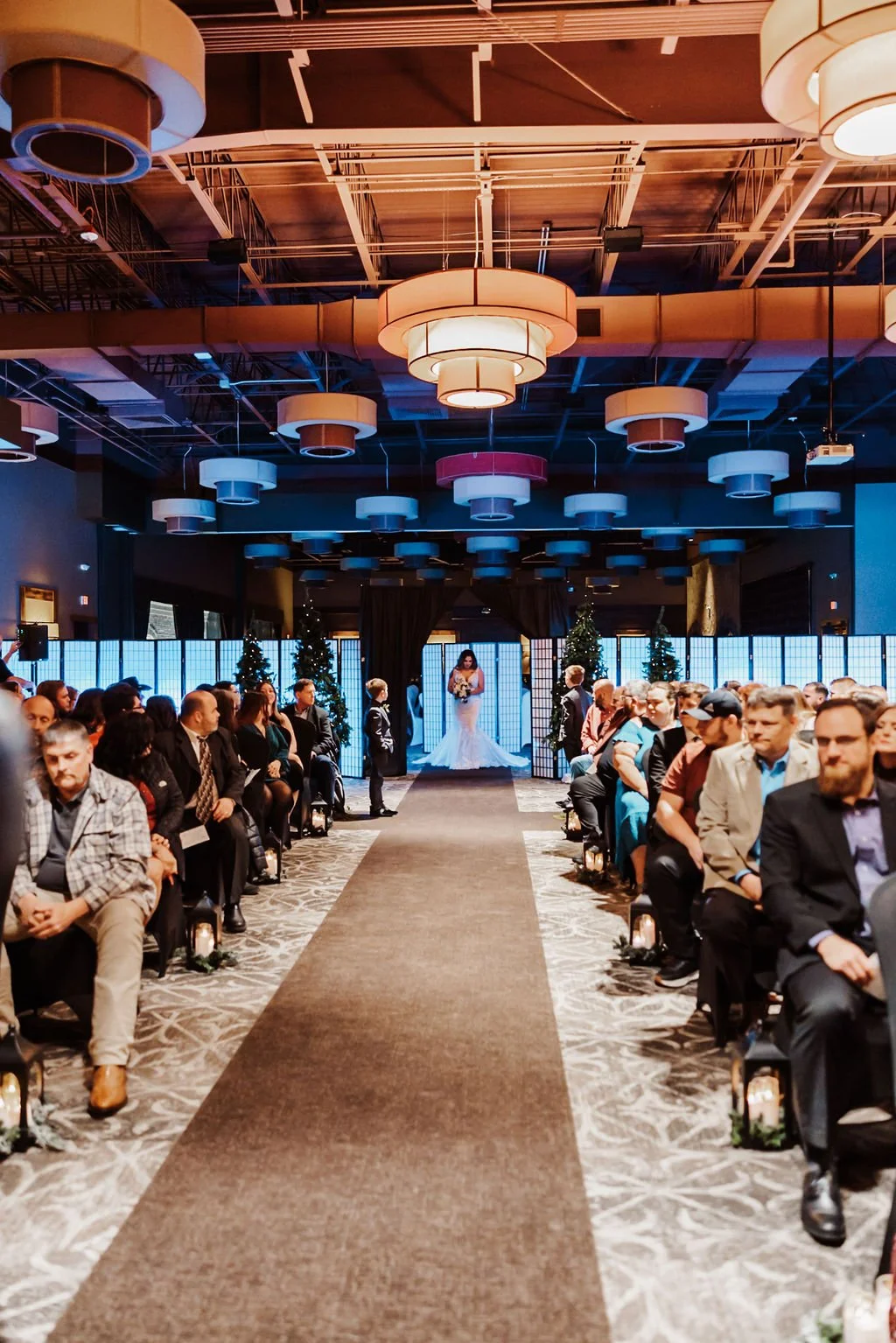 A wedding ceremony with a bride and groom standing at the altar, surrounded by seated guests, with Christmas trees and decorative screens in the background.