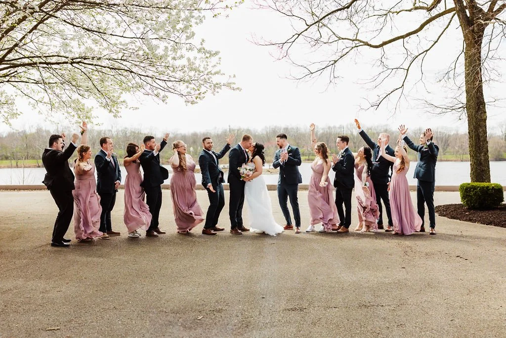 A wedding party celebrating outdoors by a lake, with the bride and groom in the center and bridesmaids and groomsmen around them, raising their hands.