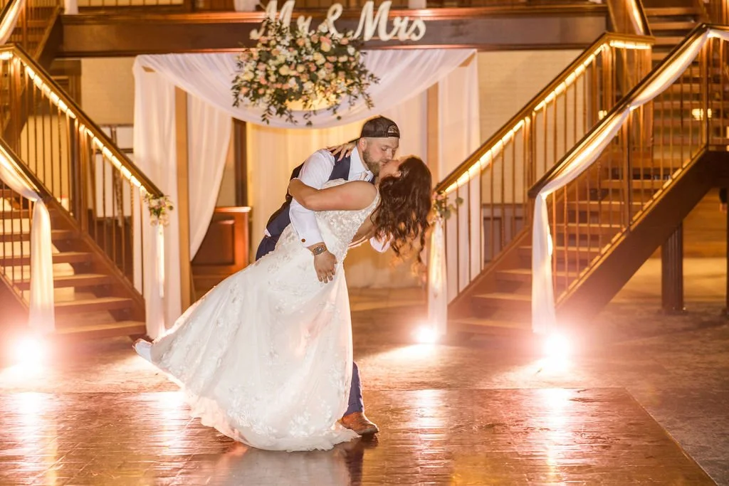 A newlywed couple sharing a kiss during their wedding reception, with the groom dipping the bride, in a decorated indoor venue with staircase and floral arrangements.