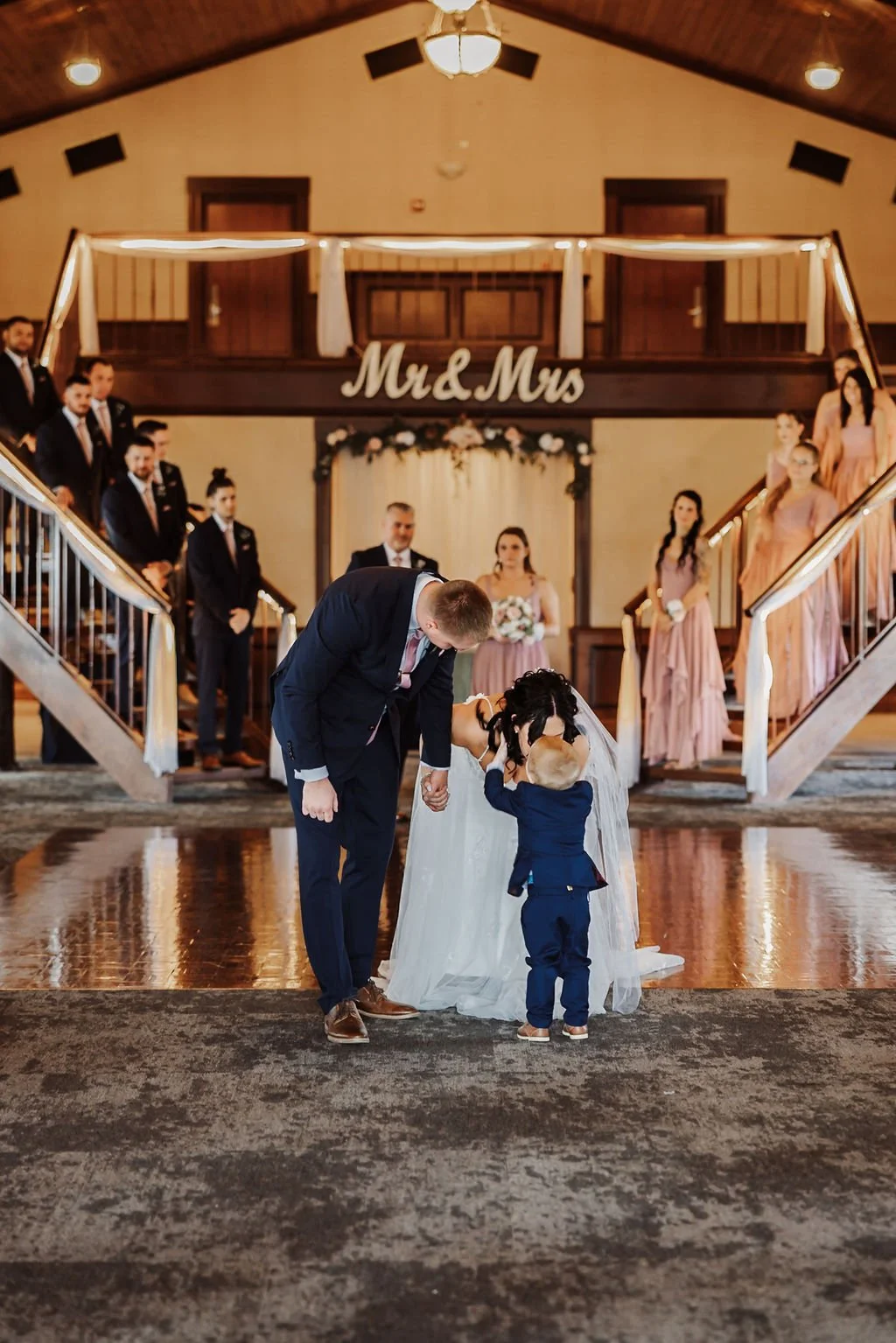 A bride and groom with a young child in a wedding ceremony. The groom is holding the child's hands, and they are kissing. The background shows wedding party members on a staircase, floral decorations, and a sign that says 'Mr & Mrs'.