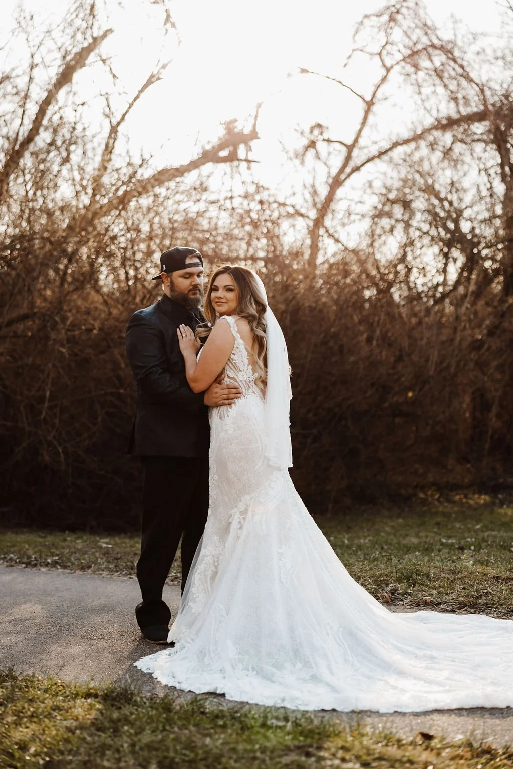 A bride and groom standing outdoors during sunset, with bare trees in the background. The bride is wearing a white lace wedding gown, and the groom is dressed in black with a backwards baseball cap. They are embracing each other, looking content and 
