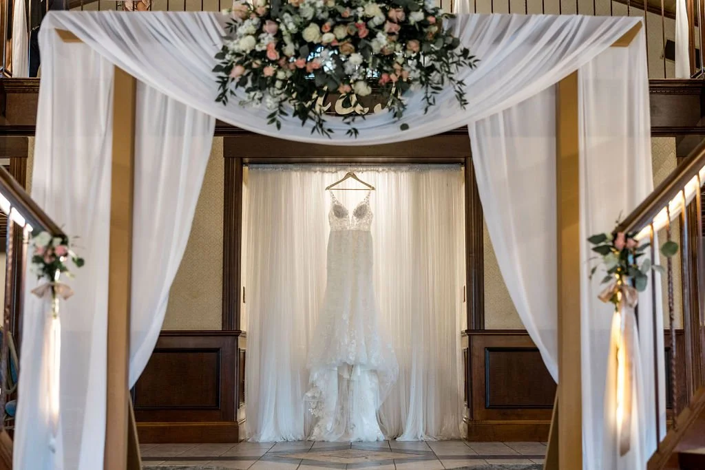 A wedding dress hanging in a bridal suite framed by white drapes and floral arrangements, with a floral Decorative arrangement at the top