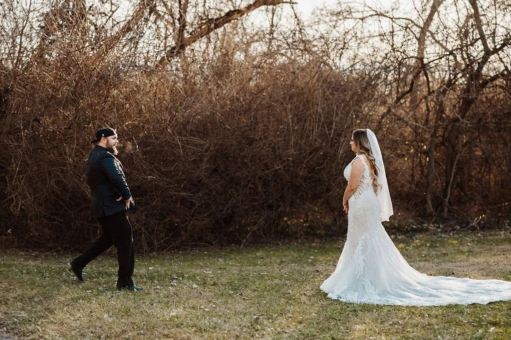 Bride in a white wedding gown and veil facing groom in a dark suit outdoors with leafless trees and bushes in the background.