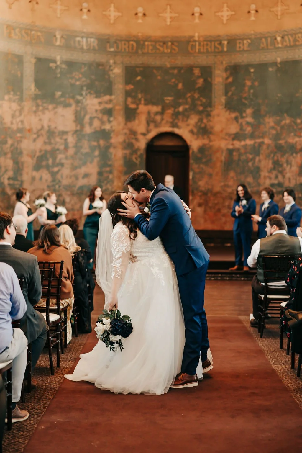 A bride and groom kiss during their wedding ceremony inside a church, with guests seated on chairs on either side and bridesmaids in teal dresses standing in the background.
