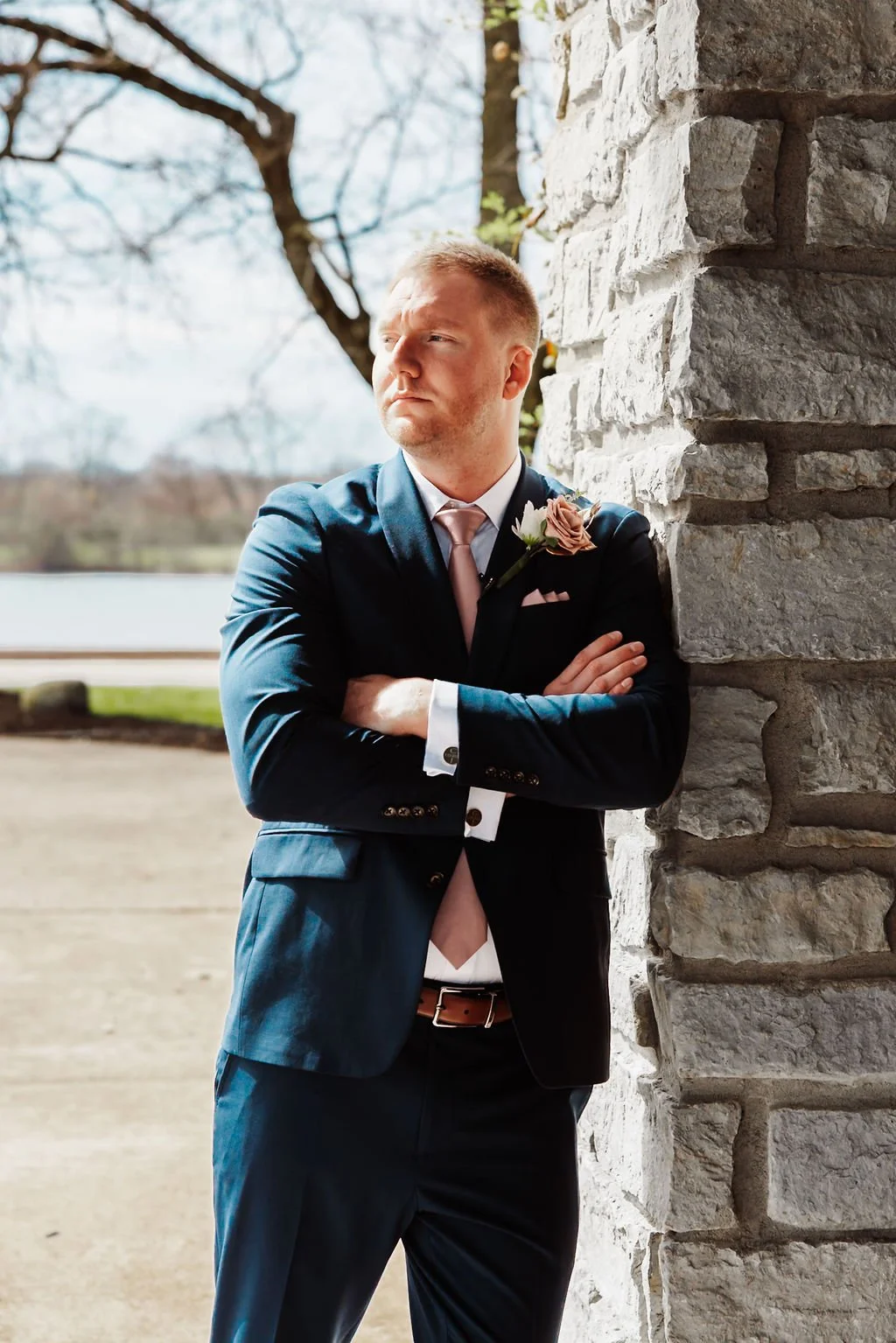 A man dressed in a dark suit and pink tie, with a boutonniere, leaning against a stone wall outdoors, looking thoughtfully into the distance with arms crossed.