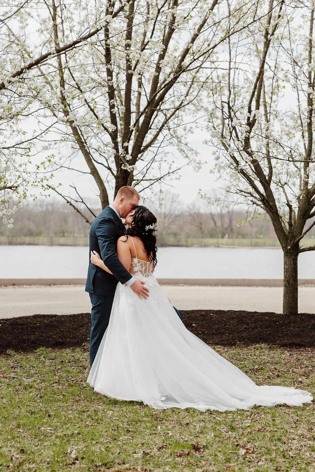 A bride and groom sharing a kiss outdoors near a lake, surrounded by trees in bloom.