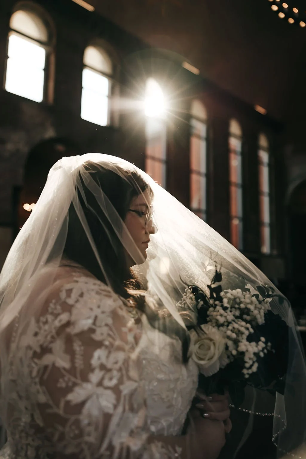 A bride in a lace wedding dress and veil holding a bouquet of white flowers inside a church with stained glass windows and sunlight streaming through.