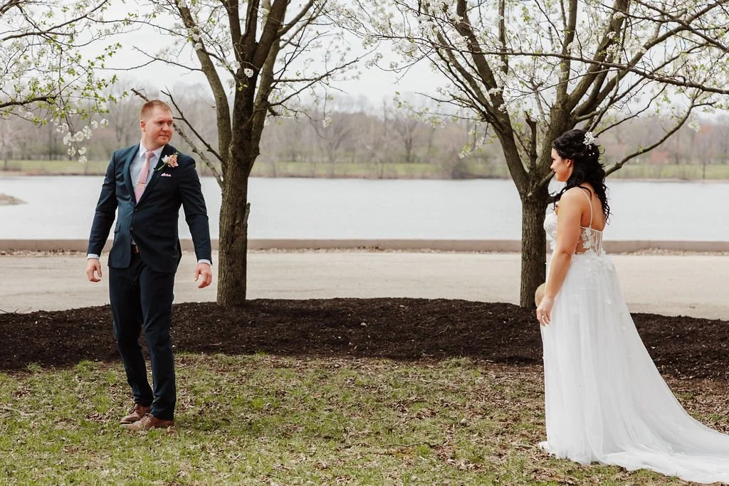 A bride and groom standing outdoors near a lake with trees in the background. The groom is in a dark suit with a pink tie, looking at the bride. The bride is in a white wedding dress with lace details, facing the groom.