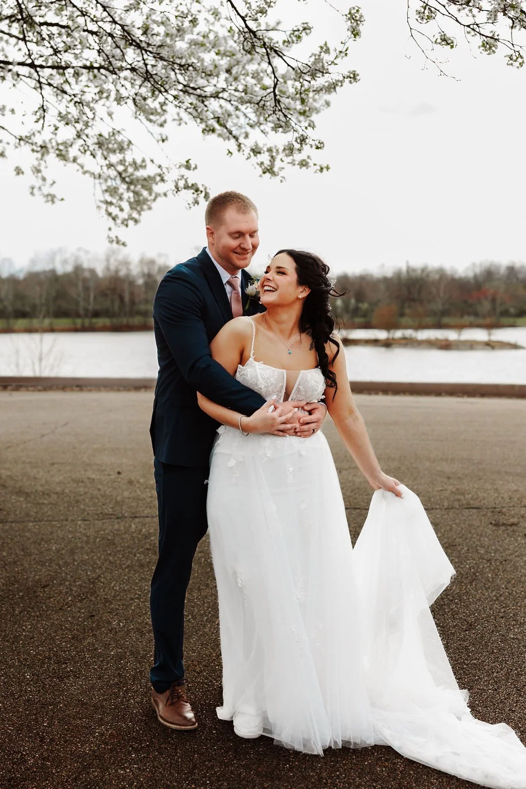 A newlywed couple stands outdoors under a tree, smiling and embracing near a lake, with trees and overcast sky in the background.