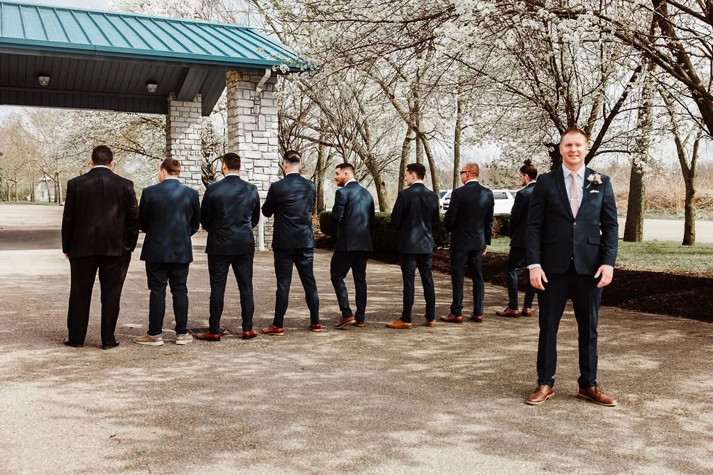 A group of men dressed in suits standing in a line outdoors, with one man standing separately and smiling at the camera.