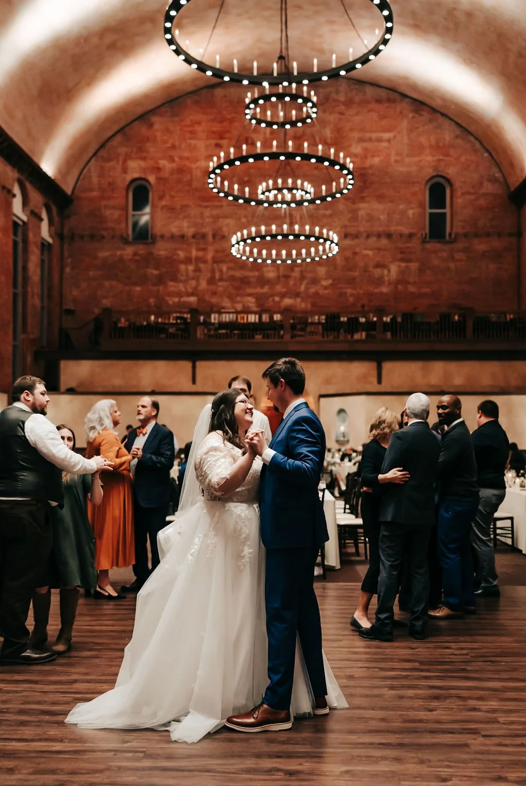 Bride and groom dancing at their wedding reception in a large, rustic hall with warm lighting and chandeliers, surrounded by guests